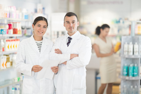 Positive man and girl pharmacists are standing in pharmacys sales hall, pharmacy staff are amicably preparing to meet chemist shop visitorsの写真素材