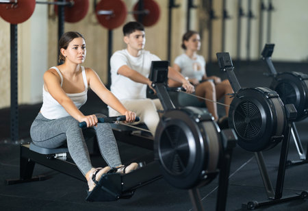Workout girl cross training exercising cardio using rowing machine with group of people in clubの写真素材