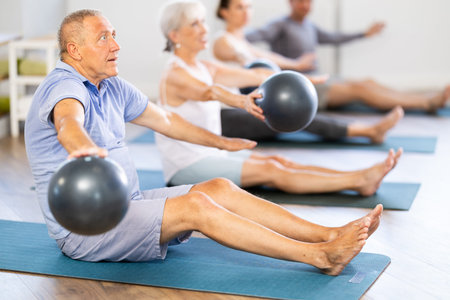 Group of mature people in sportswear exercising Pilates with soft balls while sitting on mats in...