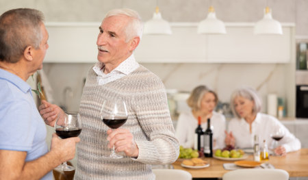 Elderly men have withdrawn from their female companions, stand with glass of wine in hands, chattingの写真素材