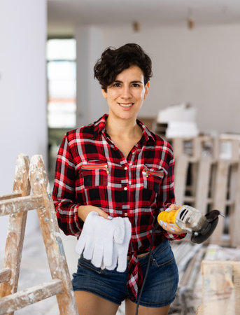 Pretty woman in a plaid shirt and short shorts poses playfully with a angle grinder in renovated roomの写真素材