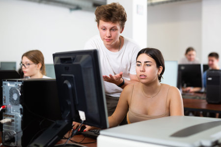 Smart teen schoolboy helping cute girl classmate sitting at computer during informatics lesson