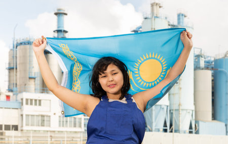 Positive girl in overalls with the Kazakhstan flag in her hands against the background of chimneys of modern factoryの写真素材
