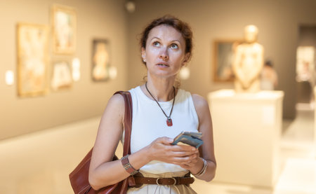 Attractive young woman in casual clothing with phone in hands looking painting in museumの写真素材