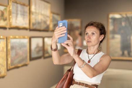 Interested woman looking closely at paintings in art gallery, using smartphone and taking photoの写真素材