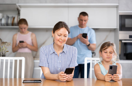 Tween girl with mother absorbedly staring at screens of smartphones in kitchenの写真素材