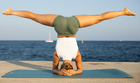 On windy day young slender woman does yoga near sea doing shirshasanaの写真素材