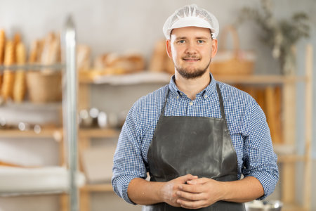 Portrait of positive baker in an apron against the background of display case with fresh buns and breadの写真素材