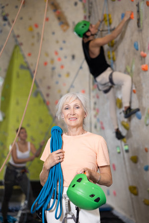 elderly positive slim woman with necessary equipment stands in gym near climbing wallの写真素材