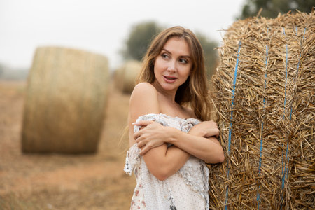 Sexy woman in white dress having picnic standing by hay bales in farm fieldの写真素材