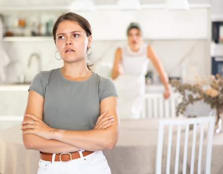 Offended girl sits at table in kitchen and listens to woman scolding herの写真素材