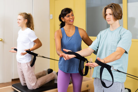 Pilates instructor helping teen boy and girl doing exercises on reformer apparatus at gym closeup