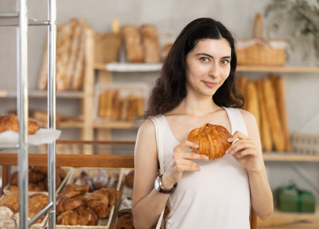 Young female customer stands with croissant in hands near window of bakery.の写真素材