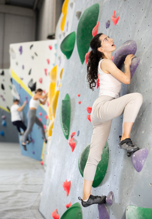 Girl is mastering climbing on training wall in gym. Woman holds on tightly to ledges and strives for top of bouldering routeの写真素材