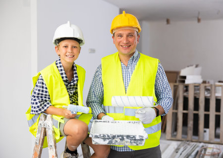 Portrait of father and son in yellow vest and protective helmet, who paint walls of room with paint...