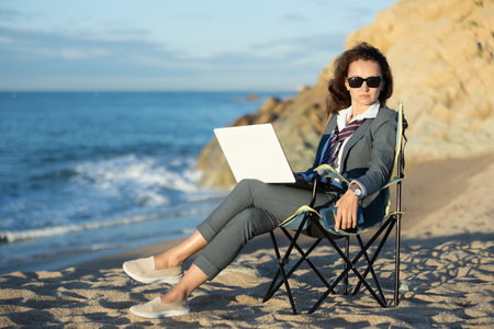 Businesswoman in sunglasses working on laptop on ocean beachの写真素材