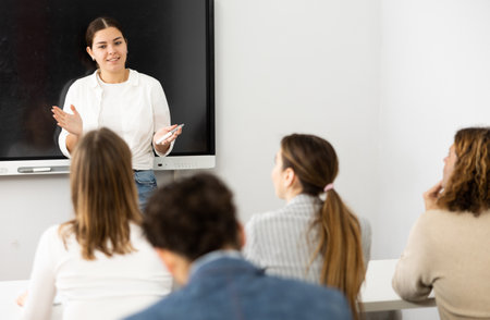Positive female trainer standing near interactive board and conducting advanced training courses to office employees sitting at desks in classroomの写真素材