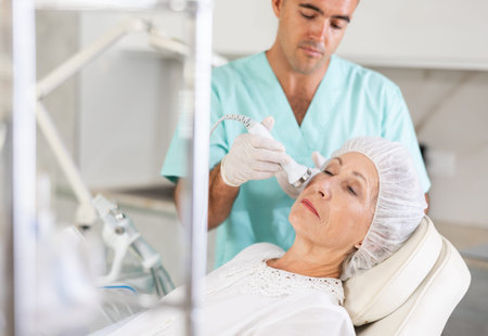 Cosmetologist performing anti-aging face hardware therapy procedure for aged female client in clinic...