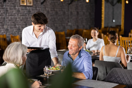 Young waiter serving ordered dishes to senior couple in restaurantの写真素材