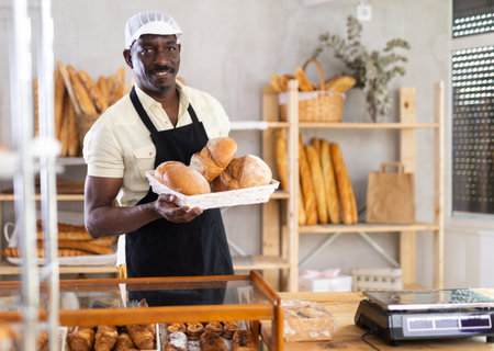 African male baker worker sells loaf of bread, shows many productsの写真素材