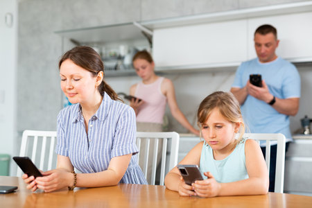 Mom sitting near kitchen table next to younger daughter with phones playing online gameの写真素材