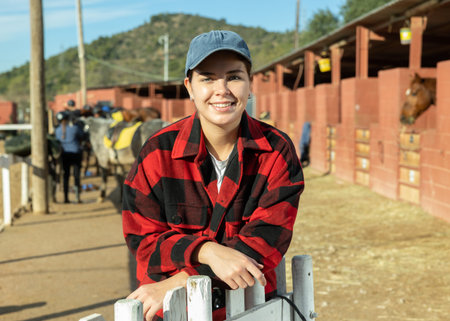 Portrait of positive smiling young woman wearing plaid shirt and posing on yard of horse stables