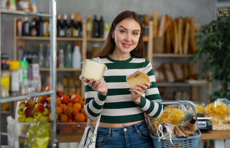 Smiling young girl with shopping basket trying to make choice holding two sorts of cheese in grocery storeの写真素材
