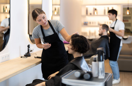 Young woman barbershop employee stands next to female client, discussing details of haircutの写真素材