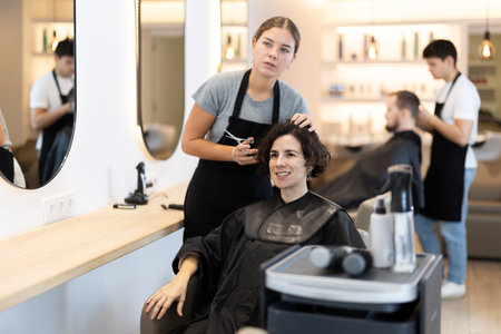Young woman barbershop employee stands next to female client, discussing details of haircutの写真素材