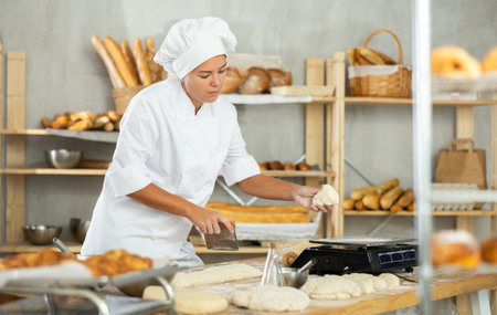 Young female baker slicing and weighting dough in bakery