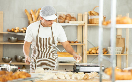Young male baker cutting and weighing dough on scales in bakehouseの写真素材