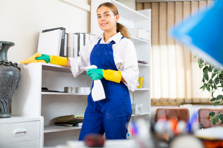 Young female janitor dusting furniture in office with cleaning equipmentsの写真素材