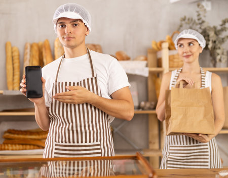 Smiling bakers showing phone and holding paper bag in bakeryの写真素材