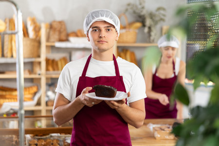 In bakery, young man seller holds piece of cake on disposable plate.の写真素材