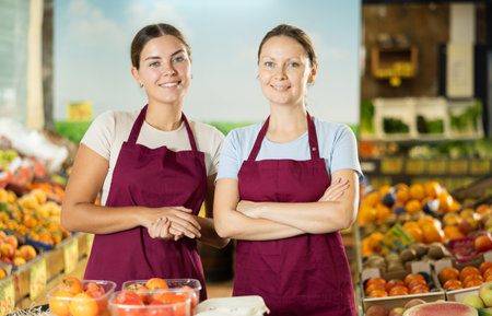 Friendly woman owner of fruit-vegetable shop standing in trading floor with girl assistantの写真素材