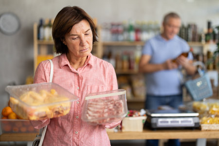 Mature woman in store took raw chicken and pork meat from shelf, choose meat, man in backgroundの写真素材