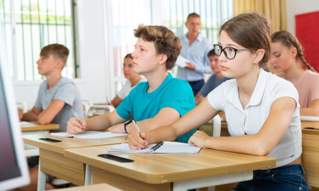 Teen girl in glasses listening to lecturer and writing in notebook in classroomの写真素材