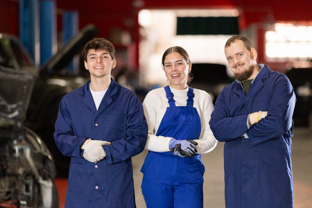 Team of auto mechanics, two men and one woman, wearing blue overalls and posing, ready to provide...