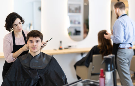 Young woman barbershop employee stands next to male client, discussing details of haircutの写真素材