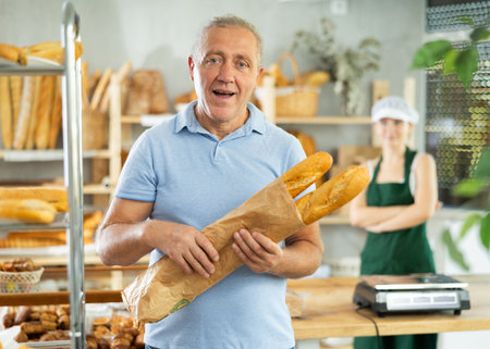 Portrait of smiling male customer holding paper bag of fresh baguettes in bakery interiorの写真素材
