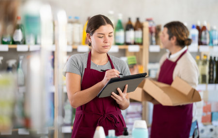 Female manager makes notes on an electronic tablet - conducts an inventory. In background, male salesperson places goods on shelvesの写真素材