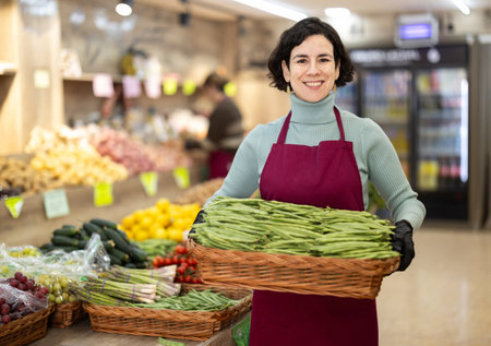 Woman salesperson carries large basket of beans in pods from warehouse to sales area in shopの写真素材