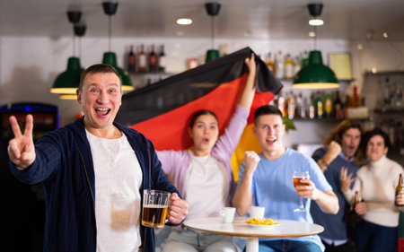 Group of young adult sports fans rooting for favorite team and waving flag of Germany while watching match together in pubの写真素材