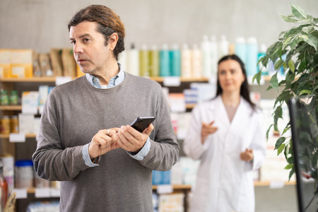 Male customer looking for something in a drugstore, checking the list on his smartphone. Female pharmacist offers medicinesの写真素材
