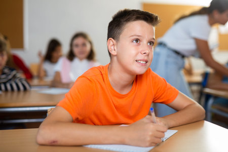 Tween boy listening to schoolteacher and writing in notebook in classroom