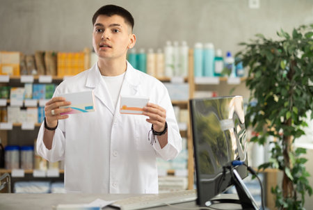 Skilled young man pharmacist standing at prescription counter and offering medicinal products to...