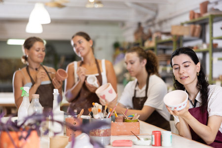 Young woman sits near table and decorates clay ceramic craft vase, plate
