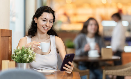 Woman holding mobile phone in her hands and drinking coffee while sitting at a tableの写真素材