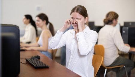 Female student rubs her tired eyes with her hands after working for long time at computer monitor in classroomの写真素材