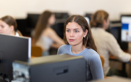 Young woman student of computer courses looks at monitor screen, types on keyboard and does workの写真素材